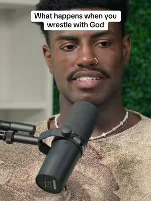A man with dark skin and a short haircut is centered in the frame, holding a microphone. A white rectangle with the text "What happens when you wrestle with God" is at the top. The man is wearing a patterned, tan-colored top and a beaded necklace. The background is a green, blurred texture.