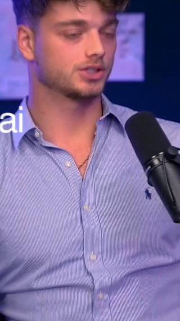 A man with short, styled hair and a light beard is centered in a medium shot, wearing a light blue, button-down shirt with vertical stripes. He is speaking, and a black microphone is positioned close to his mouth. The background includes a hint of a dark blue shade behind the man's head.