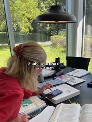 A woman is seated at a table, her back mostly to the camera, studying with books and a phone. The scene is indoors, with a bright window showing a green outdoor setting. The woman is wearing a red top and has her blonde hair up, and there is text overlaid on the image.