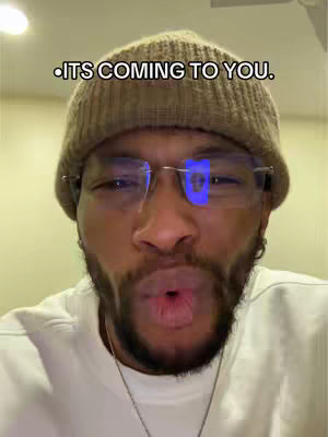A selfie-style shot of a Black man with glasses and a brown beanie, looking directly at the camera with a slight smirk and pursed lips. The background is a simple, out-of-focus interior. The overlayed text reads 'ITS COMING TO YOU.'