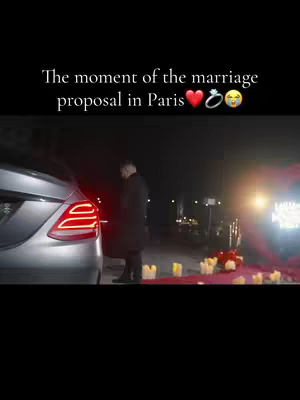 A dark, vertical frame captures a nighttime scene centered on a silver car with its brake lights on. A man in a dark coat stands near the car, facing away from the camera. In front of the car, a red carpet is laid out, with candles and possibly flowers placed along it. The background is dim, with out-of-focus lights suggesting a city environment.