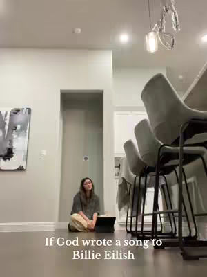 A young woman sits cross-legged on a hardwood floor, facing towards the camera. She's dressed in casual clothing against a minimalist background. The setting is a modern home, with a sleek kitchen island and chairs visible.