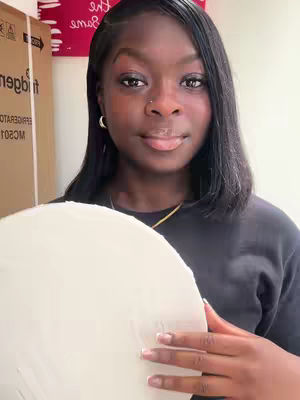 A woman of color holds a round, off-white canvas. She faces the camera with a slight smile. Behind her are some boxes and a wall.