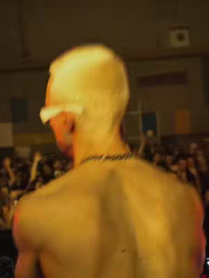 The cover image shows a man's upper back and head, facing away from the camera, in front of a crowd. He has short, possibly bleached, hair and is wearing earrings. The lighting suggests a stage environment. The depth of field is shallow, blurring the crowd in the background.