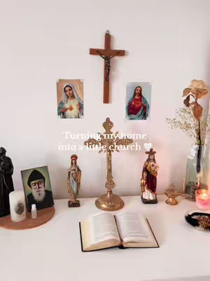 An interior shot featuring a white surface with various religious artifacts and decor. A large wooden cross is mounted on the wall above several small statues. On the surface are an open book, a small golden cross, a candle, and other religious images.