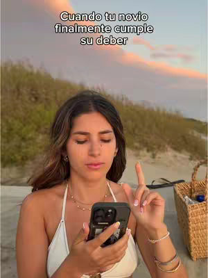 A woman with long brown hair is holding a black smartphone in her left hand and pointing her right index finger upwards. She is wearing a white top and multiple bracelets. The background is a blurred beach scene with a sunset.