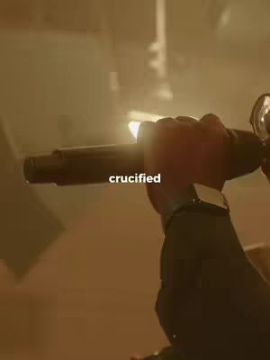 The image is a close-up shot of a person's hand holding what appears to be a cross in a dimly lit, warm-toned environment. The hand is the primary focus, with a watch visible on the wrist. The background is blurred, with a light source creating a hazy effect, making it difficult to discern the setting. Text overlayed on the image reads "crucified."