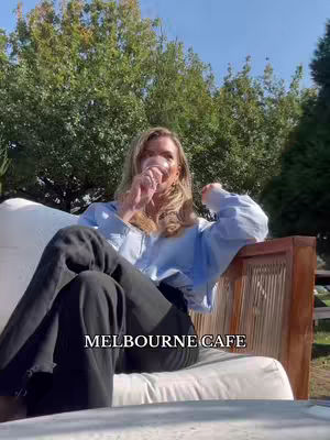A woman with blonde hair is seated on a cream-colored lounge chair in an outdoor setting. She is holding a glass to her lips, likely a drink. The background features lush green trees under a bright sky. The text overlay says, 'MELBOURNE CAFE'.