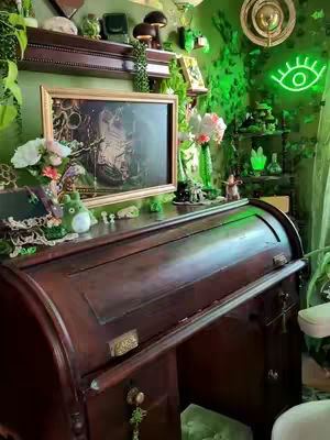 The image showcases a dark wooden roll-top desk in a room decorated with a fantasy theme. The room has green walls and various trinkets on shelves and the desk surface. A framed picture is displayed on the wall above the desk.