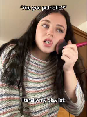 A woman with dark, long hair is applying makeup with a blush brush while looking off-camera. Text overlays read, 'Are you patriotic?' and 'literally my playlist:'. The background appears to be a room.