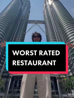 A person stands centered in the frame, arms outstretched, with the Petronas Towers in the background. A black rectangle with white text overlayed on the lower portion of the central person. The shot is taken on a clear day with a bright blue sky, with the towers appearing to rise sharply behind the person .