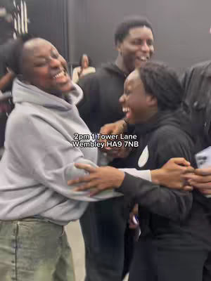 A group of young people are embracing and smiling. Two of them are in the foreground laughing, with a third person in the background. The scene is brightly lit, with a neutral wall behind them.