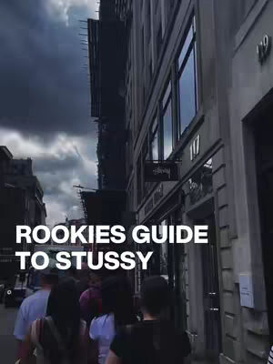 An exterior shot of a Stussy store with "ROOKIES GUIDE TO STUSSY" text overlayed. The facade of the building is visible, along with the Stussy sign. People are seen walking in front of the store.
