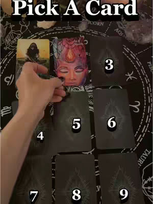 A hand is reaching towards a deck of tarot cards, with various cards laid out in a spread. The layout includes some face-up cards and numbers indicating positions. The background features a dark textile with astrological symbols.