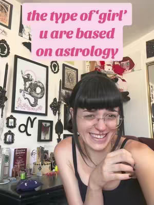 A woman with dark hair and glasses smiles, positioned centrally. A pink text overlay is above her head on a wall covered with various framed artworks.