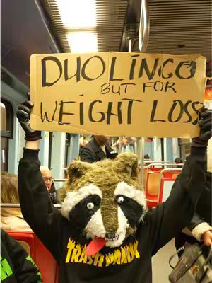 A person dressed in a raccoon mask and a hoodie that says "TRASH PANDA" holds a cardboard sign that reads "DUOLINGO BUT FOR WEIGHT LOSS," standing in a subway or transit setting. The background shows seats and other passengers.