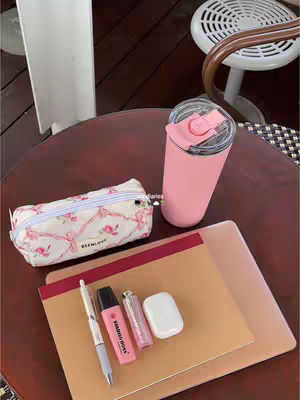 The image showcases a pink and white aesthetic. Centered on a round, brown table are several items: a pink tumbler with a straw, a pencil case with a floral design, a stack of notebooks, and a pen, highlighter, and wireless earbuds. The composition makes use of a warm color palette and organized arrangement, suggesting a study or workspace setting.