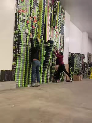 Two people are shown trying to reach the top of a wall entirely composed of boxes, likely storage or transport boxes. The boxes vary in color, mainly green, black, and white, arranged in a seemingly random but organized fashion. The lighting appears to be from overhead sources, casting shadows and highlighting the textures and colors of the boxes. Both people have their backs to the camera, with one wearing a maroon-colored shirt, while the other has a black jacket.