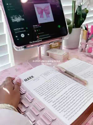 The image shows a desk setup with a Bible open and a tablet displaying a music app. The scene is bathed in pink hues. White blinds are visible in the background. The bible text is MARK.