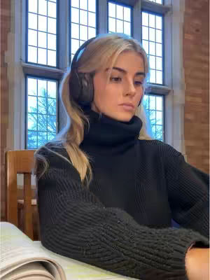 A young woman with long blonde hair and a black turtleneck sweater is seated indoors. She is wearing headphones. A large window with a grid pattern and a wooden chair are in the background. The woman is looking pensively to the side, with a book open in front of her.