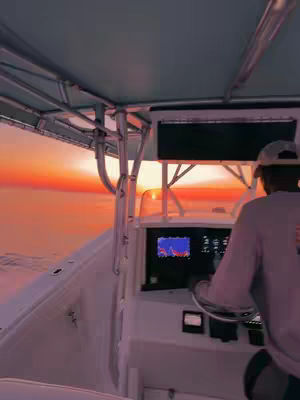 A person is piloting a boat at sunset. The image is taken from inside the boat cabin, showing the dashboard and the driver with the view of the colorful sky and water through the front. The sunset paints the sky shades of orange and pink.
