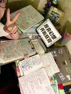 A top-down view of a cluttered desk with open notebooks, a tablet displaying Chinese characters, and a person's hand making a peace sign. The scene suggests a study environment, possibly for online content creation.