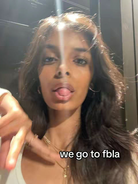 A selfie-style shot from a slightly low angle. The creator, a young woman with long, dark hair, is the central focus. She has a direct gaze, and her tongue is sticking out slightly. The background is blurred, likely indoors.