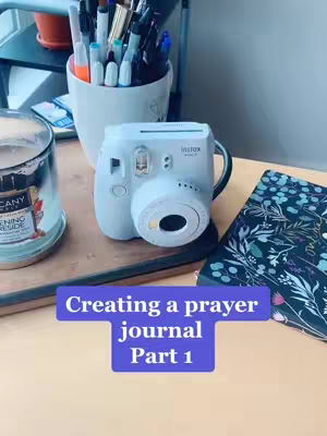 A close-up shot of a desk area featuring a white Instax camera in the center. To the left is a candle, and to the right, a notebook with floral designs. A cup filled with pens is also visible, and the image has a soft, natural lighting.