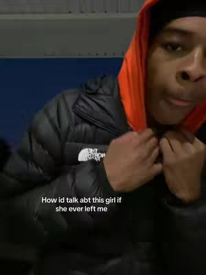 A person wearing a black puffer jacket with an orange hoodie underneath, against a dark, possibly outdoor, background. The person is looking down and holding the jacket near the chest, with text overlaying the image.