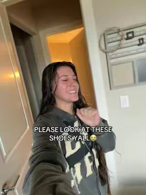 A woman with long dark hair smiles, wearing a gray zip-up hoodie with white text. Behind her are a white door and a hallway with a yellow-lit room. Text overlayed says: 'PLEASE LOOK AT THESE SHOES YALL' with a smiley face.