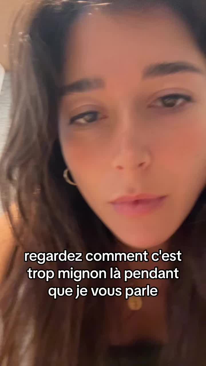 A selfie-style shot of a woman's face, with text overlayed at the bottom. The woman has dark hair and is looking towards the camera. The background is out of focus. The text overlay is in white and appears to be a conversation.
