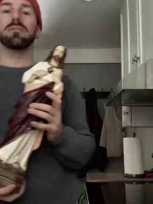 A man with a red beanie is holding a statue of Jesus in a kitchen setting. The statue is white and brown and has a detailed face. The man's face is visible, looking at the camera. Behind him are kitchen cabinets and other appliances.