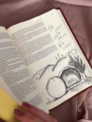 An open bible resting on a pinkish surface. The right page has an illustration of an empty tomb with three crosses on a hill. Calligraphic text is written on the right page as well. A hand is visible in the bottom left corner.