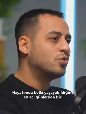 A medium shot shows a man in the foreground, facing slightly to his right and speaking. He has short, dark hair and is wearing a black sweater. A microphone is partially visible to his right. Text overlayed on the bottom of the image reads, in Turkish, "Hayatımda belki yaşayabildiğim en acı günlerden biri" (One of the most painful days of my life). The background is blurred, with a glimpse of a light-blue wall.