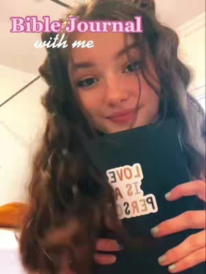 A young woman with long, wavy brown hair is centered in the frame, holding a black book close to her chest. The book has a white sticker on it with the words in a mirrored format. The background appears to be a room with some minimal decor. Overlayed text at the top reads "Bible Journal with me". The overall tone is warm and inviting.