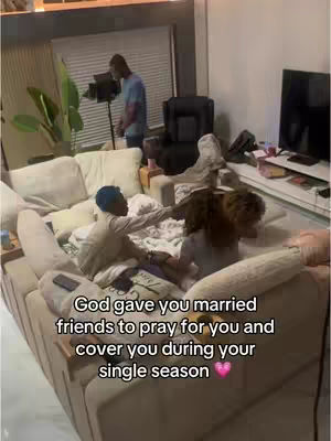 The image shows a room with light-colored walls and a large, beige sectional sofa. Two people are sitting on the sofa, one is leaning over the other, and appear to be embracing. A person stands in the background near what looks like a lighting setup. The text overlay reads a message of faith and friendship.