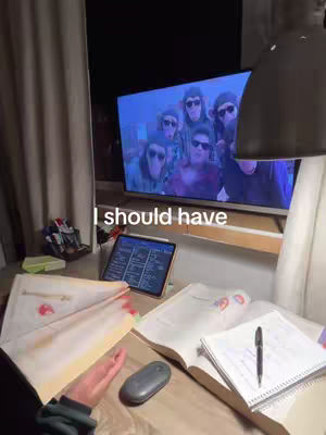 A cluttered desk is in the foreground, with papers and a notepad. Behind, a large TV shows a group of people in sunglasses. Text overlayed reads 'I should have'.
