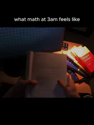 A dimly lit scene features a person's hands holding open a book, the pages filled with text. In the background, a computer screen displays a grid pattern, alongside a collection of study books stacked on a desk illuminated by warm light, suggesting late-night study. A remote control lies nearby, enhancing the impression of a study environment, the overall atmosphere is focused on the academic study.