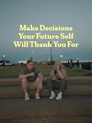Two men are seated side by side outdoors. The image appears to be taken at dusk or dawn, with a muted sky and a glimpse of city lights in the background. The men are positioned on what appears to be a low wall or curb, and the image has a soft, slightly desaturated color palette.
