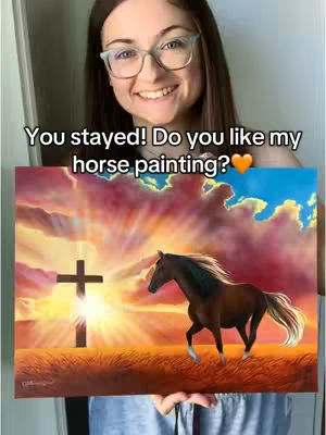 A woman smiling and holding up a painting of a horse running towards a sunset, with a cross silhouette in the background. The painting sits in the foreground, the woman in the background.