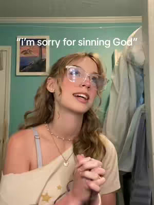 A young woman with long blonde hair and glasses is the primary focus, standing in front of a light turquoise wall. She is wearing a light-colored top and a necklace, with her hands clasped. Above her, a text overlay reads, "'I'm sorry for sinning God'". A framed painting hangs on the wall, while a white coat is hanging on the right.
