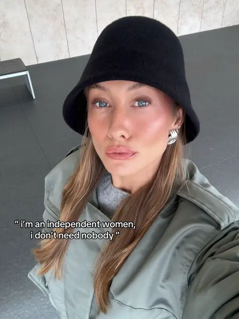 A selfie-style image of a woman with light skin and blue eyes, wearing a black hat and a green jacket. She has full lips and is looking directly at the camera. Text overlayed on the image reads, "I'm an independent woman, I don't need nobody."