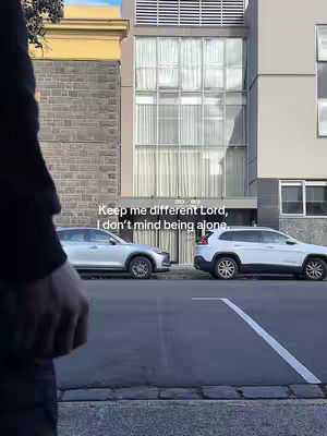 A person's silhouette is seen from the waist up. The person is on the left, with their back to the camera, in front of a building with a modern design. Two cars are parked on the street in front of the building.