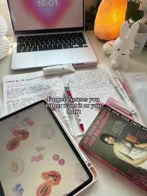 The image is a flat lay of study items on a white desk. A opened laptop is in the upper part of the frame. Below the laptop is an open notebook with writing, a pen, and a book titled 'My Year of Rest and Relaxation'. In the bottom left corner, a tablet with illustrations on screen and a pen are placed. Additionally, there is a decorative white bunny, a salt lamp, and other study items.