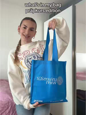 A young woman with long blonde hair in a braid holds up a blue tote bag. The bag features a logo and text, and she smiles at the camera. She is wearing a white sweatshirt and standing in front of a white sliding door and a pink sheet.