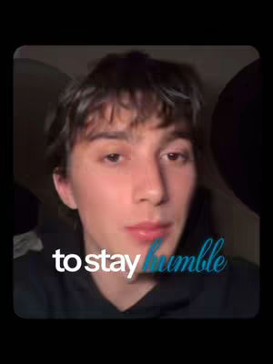 The cover image is a close-up shot of a young man's face. The focus is on his upper body, with a dark hoodie and the background being out of focus, also dark, suggesting a dimly lit setting. The man has short, dark hair, fair skin, and is looking at the camera. Overlayed in a stylized cursive font is the text 'to stay humble' across his face. The composition is simple, drawing attention to the man and the message.