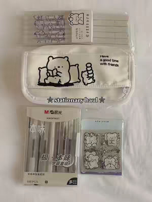 The image is a top-down shot of various stationery items arranged on a light-colored surface. In the center is a white pencil case with a cartoon bear illustration and text. Above it, there are clear plastic packets with pens and pencils. Below the pencil case, there are boxes of pens and stickers. The overall aesthetic is clean and organized, with a soft color palette.