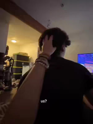 A person is seen from the back, with their arm reaching forward to touch the back of another person's head. The setting appears to be an interior space, possibly a living room, with warm lighting and a hint of a television screen in the background.