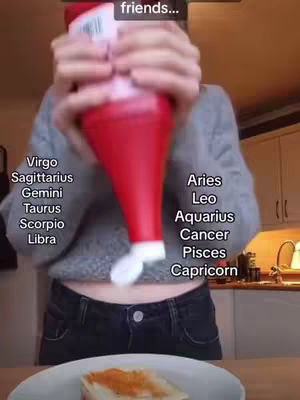 The cover image is a selfie-style shot where a person holds a red, squeeze bottle of sauce directly in front of their face. The bottle obstructs the face, but the person’s midriff and a plate with food are visible below. Text overlays on either side of the bottle lists zodiac signs, with the left side showing 'Virgo, Sagittarius, Gemini, Taurus, Scorpio, Libra' and the right side listing 'Aries, Leo, Aquarius, Cancer, Pisces, Capricorn'. The background is a kitchen, with cabinets and countertops indirectly visible.