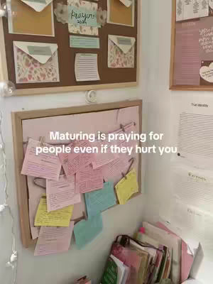 A wall with multiple framed bulletin boards. The top board is brown and displays colorful envelopes, handwritten notes, and a 'Prayer 2024' sign. The lower board shows numerous handwritten notes clipped onto strings. The wall is light, and a stack of notebooks sits below the lower board.